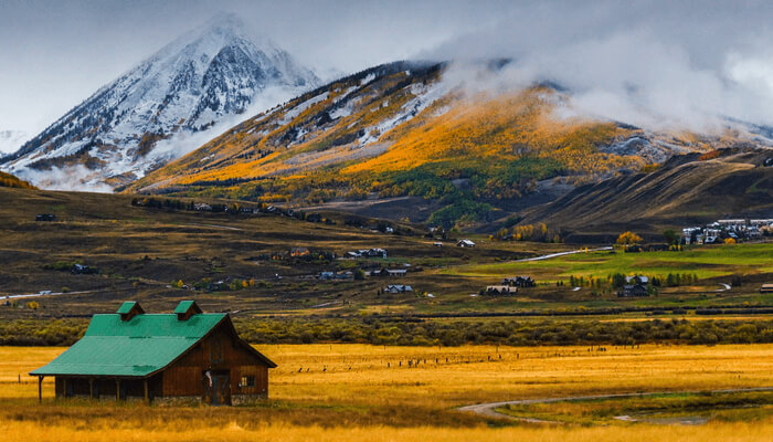 Image of a wooden cabin with a green roof with stunning mountains and clouds in the background.
