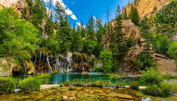 Image of the iconic Hanging Lake in Glenwood Springs, Colorado- The water is vivid blue and there are trees and rocky walls surrounding this stunning area.
