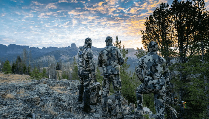 Image of three individuals in hunting gear. They are standing as they look over the mountains and soak in the mountain views.