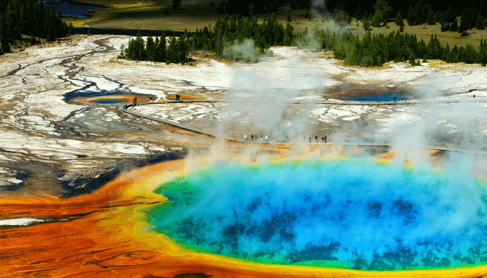 Image of Yellowstone National Park's Old Faithful- there are stunning colors of blue, green, yellow, and orange as people walk around the thermal water on the sightseeing path.