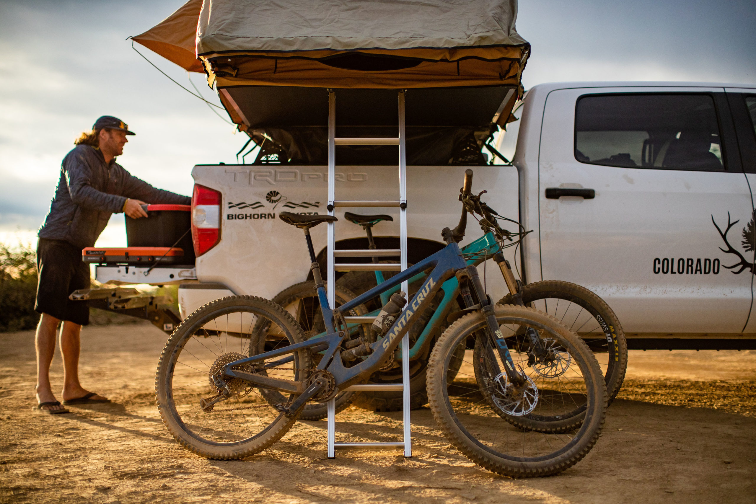 Image of a man unloading his Colorado Overlander truck preparing for his bike ride. There are two bikes leaning against the ladder to the roof nest tent.